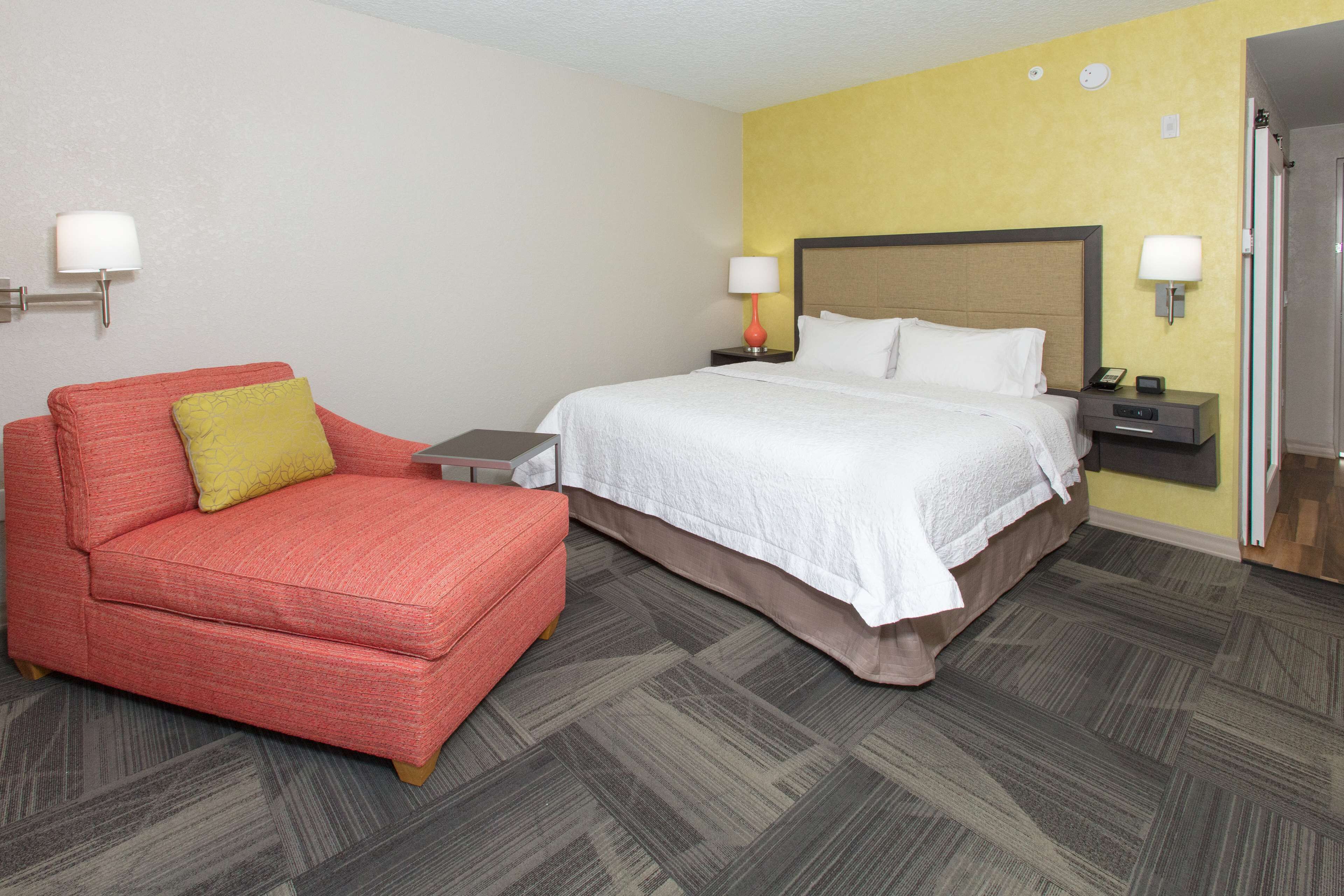 A hotel room with a bed, a red armchair, a nightstand, and a wall-mounted lamp.