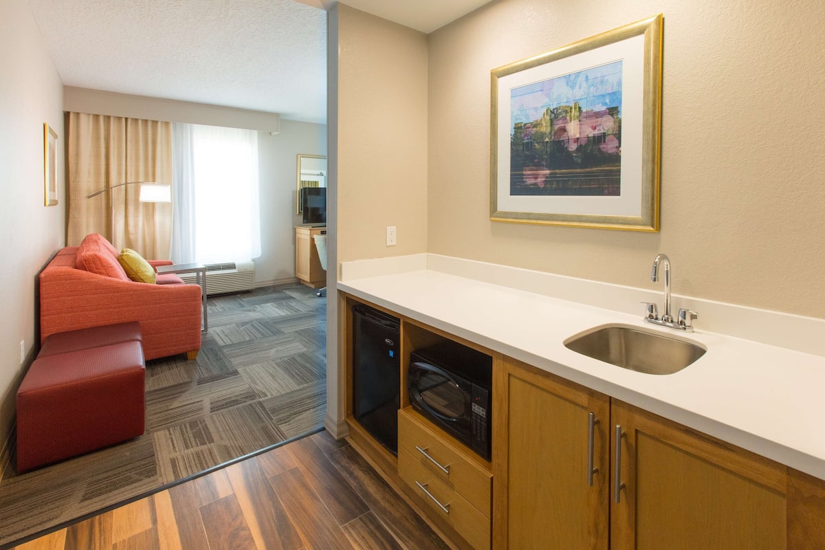 Habitación de hotel compacta con kitchenette, un sofá rojo, una pequeña mesa de comedor y un cuadro en la pared.