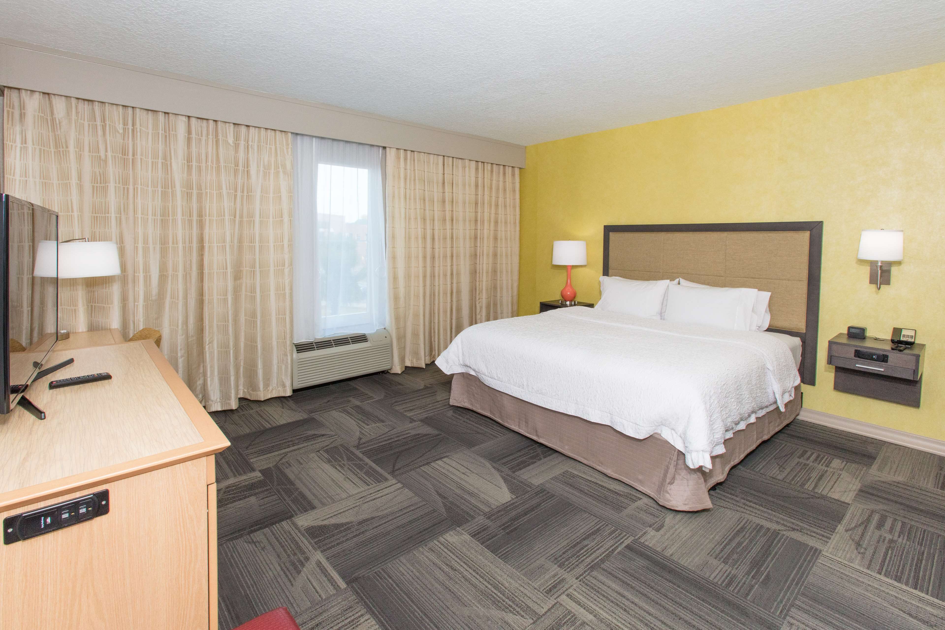 A hotel room with a bed, bedside tables, a desk with a chair, and a large window with curtains.