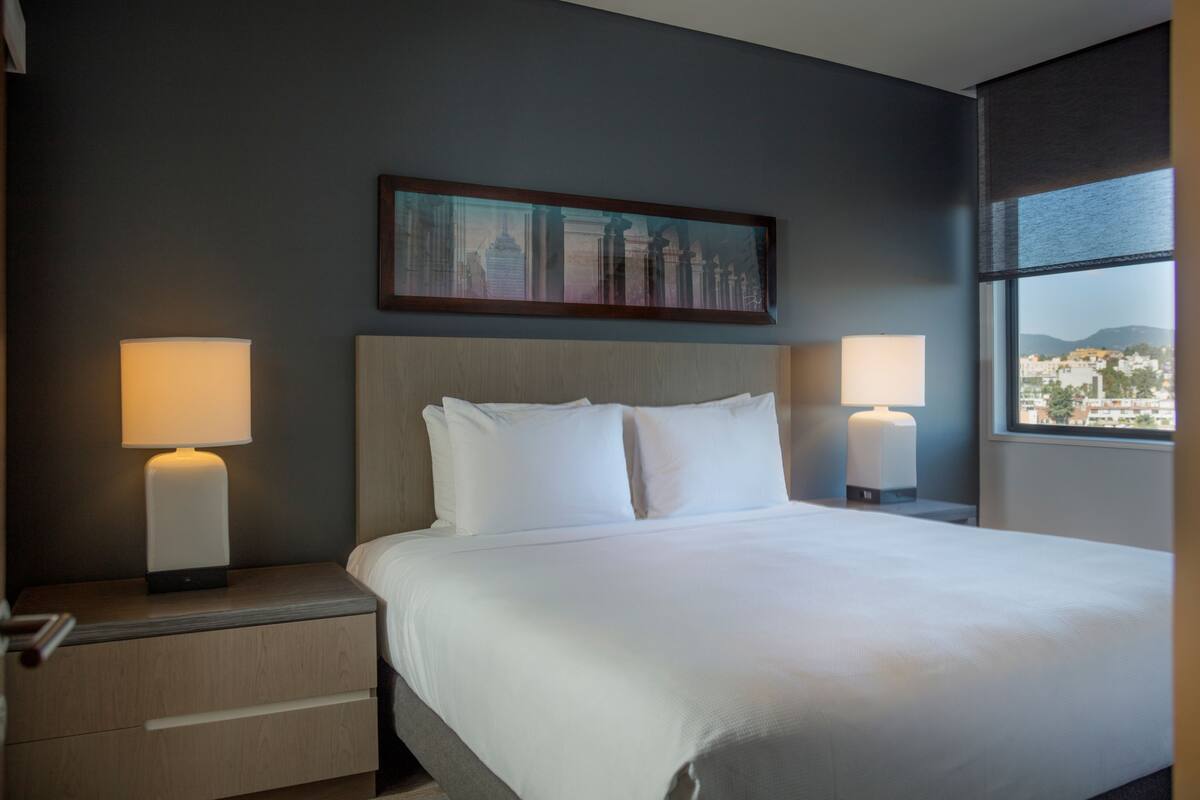 A hotel room with a large bed, two bedside lamps, a nightstand with a drawer, and a cityscape painting on the wall.