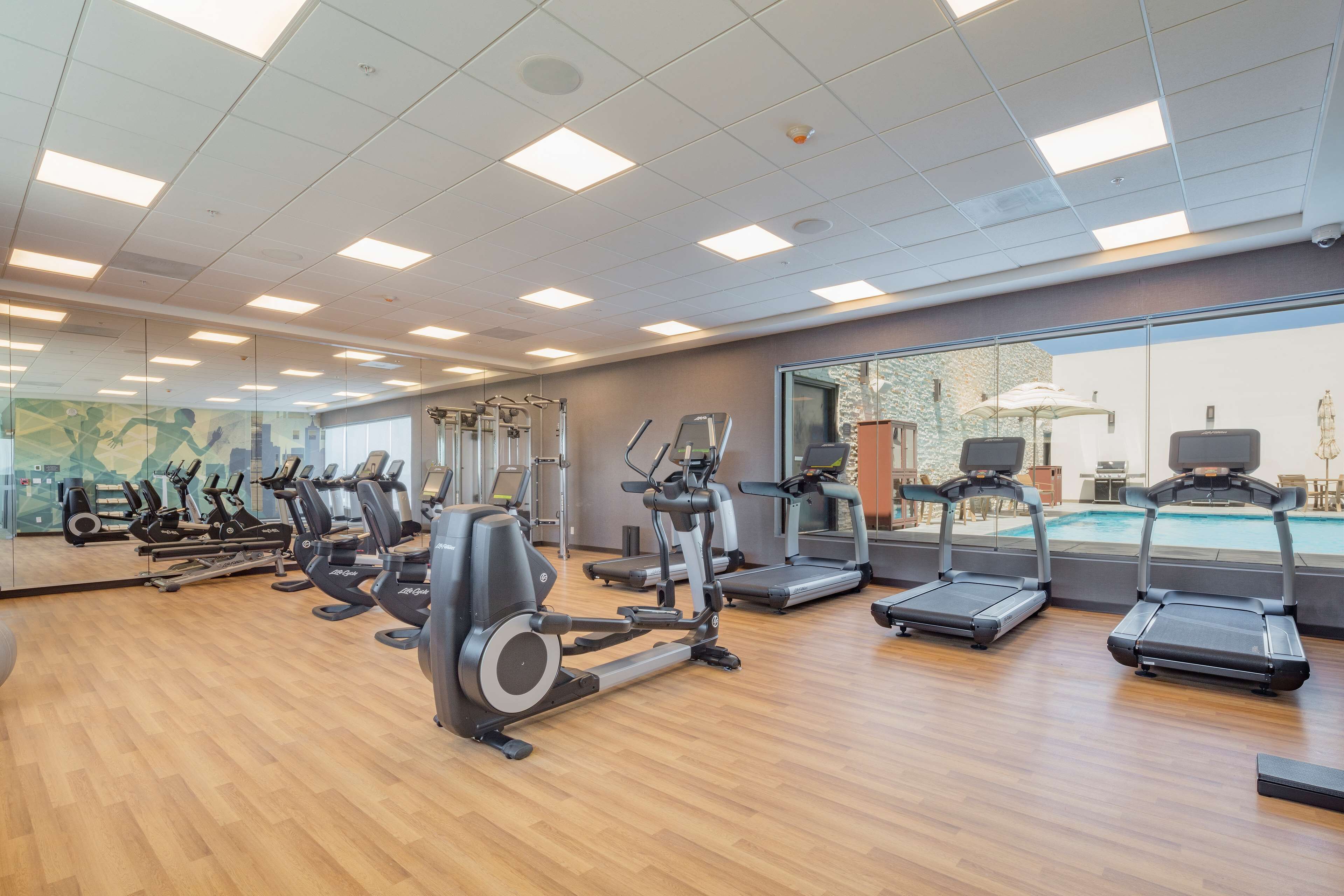 Un gimnasio con diversas máquinas de cardio, una piscina visible a través de un muro de cristal y suelo de madera.
