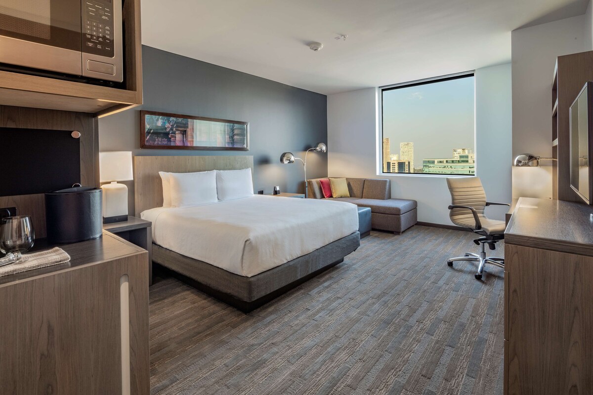 A modern hotel room with a large bed, a desk, a chair, a window with a city view, and a microwave.