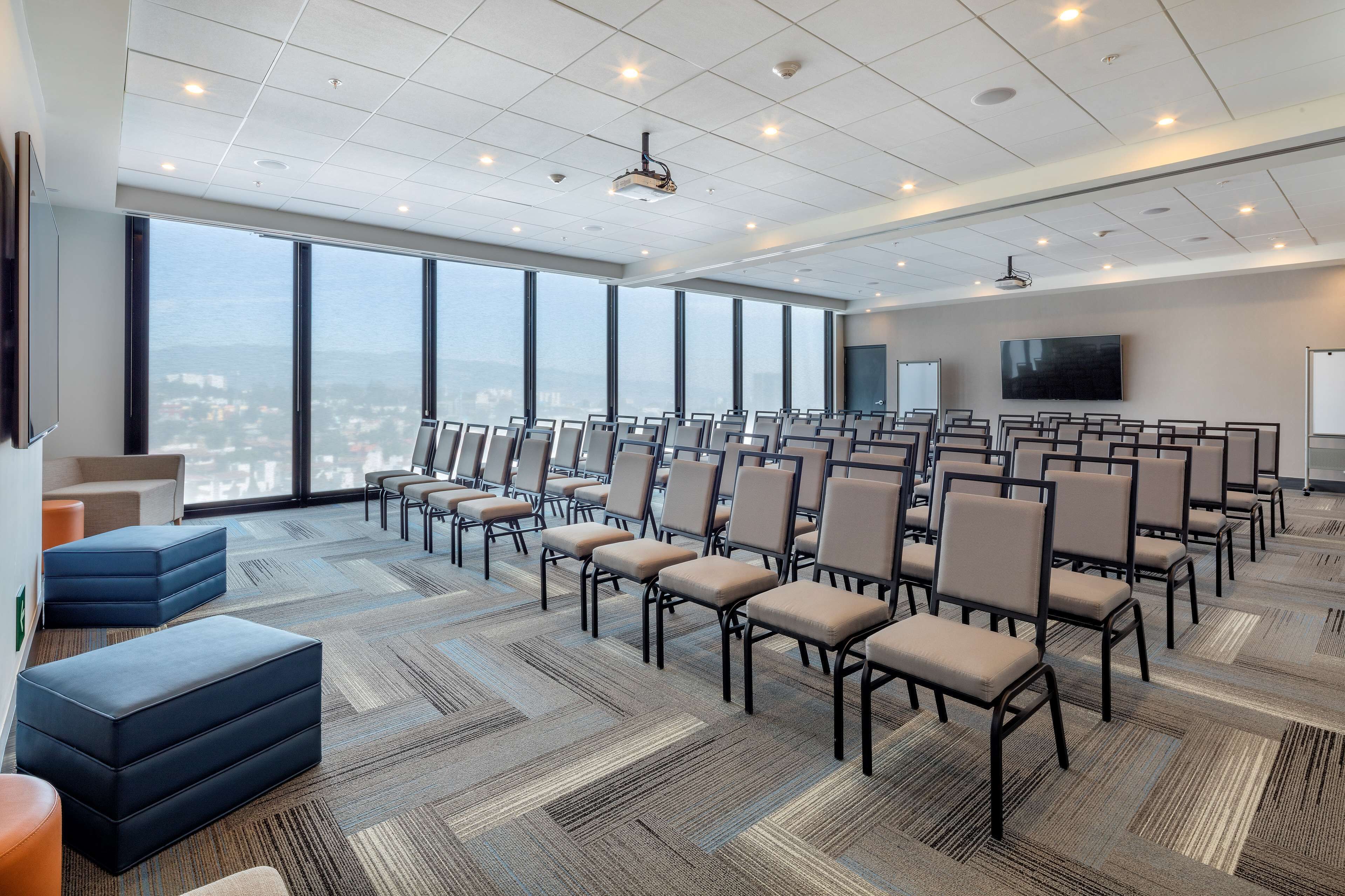 Un salón de conferencias con filas de sillas, un proyector y un amplio ventanal con vista a la ciudad.