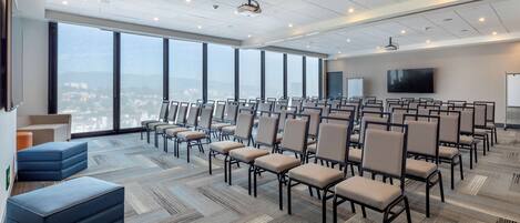 Un salón de conferencias con filas de sillas, un proyector y un amplio ventanal con vista a la ciudad.
