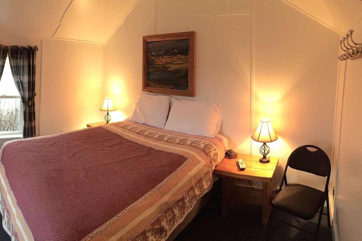A hotel room with a bed, two bedside tables with lamps, a chair, a television on a stand, a window with curtains, and a painting on the wall.