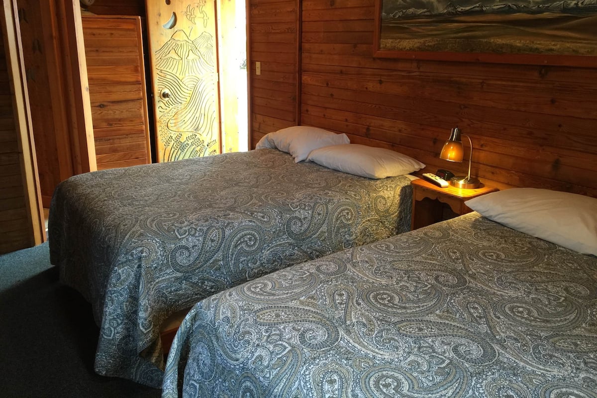 A wooden cabin room with two beds, a painting on the wall, and a small table with a lamp.