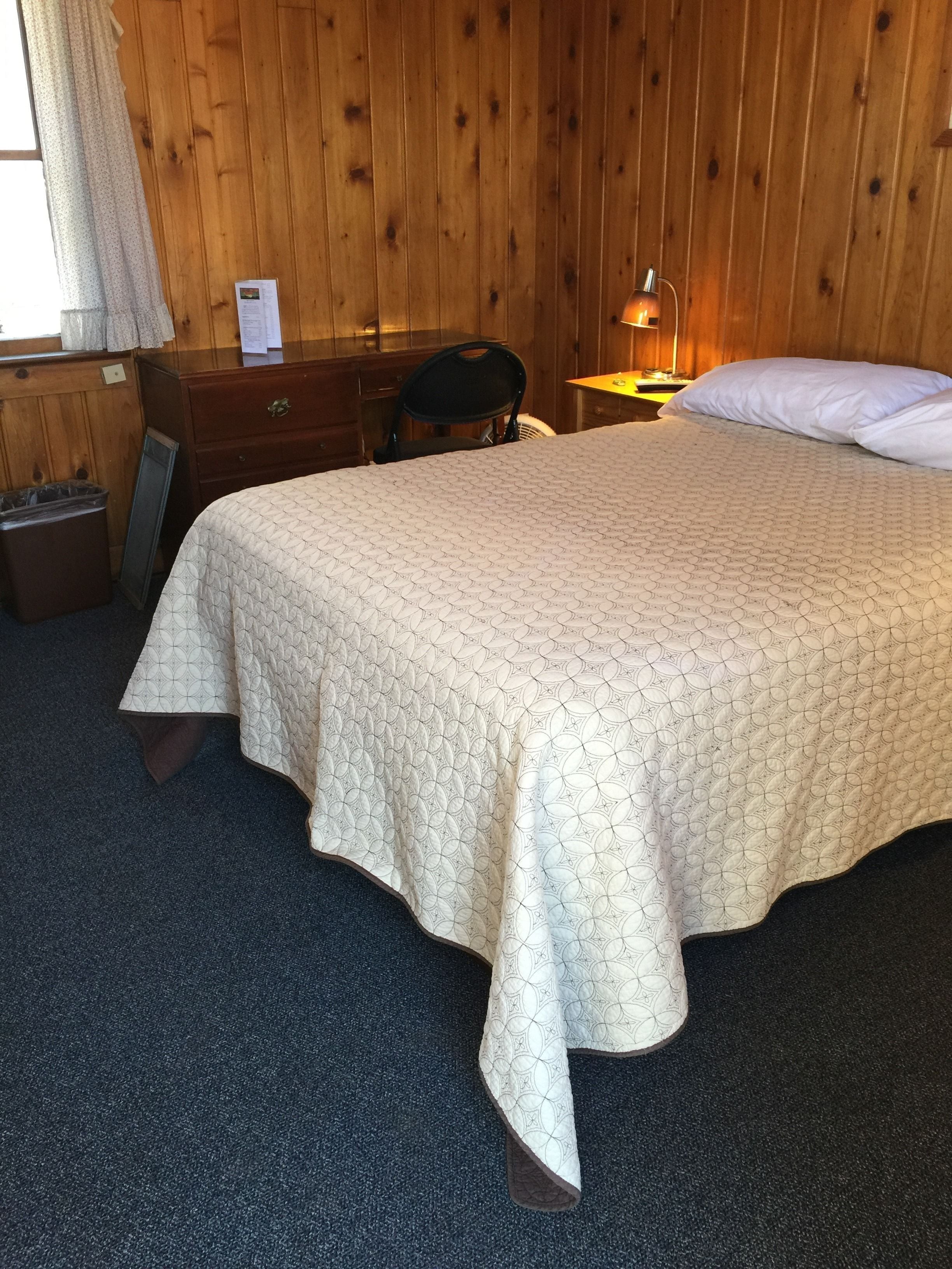 Ein ordentlich bezogenes Bett mit einem gemusterten Überwurf, ein Nachttisch mit Lampe, ein Sessel und ein kleiner Mülleimer in einem Zimmer mit Holzwänden.