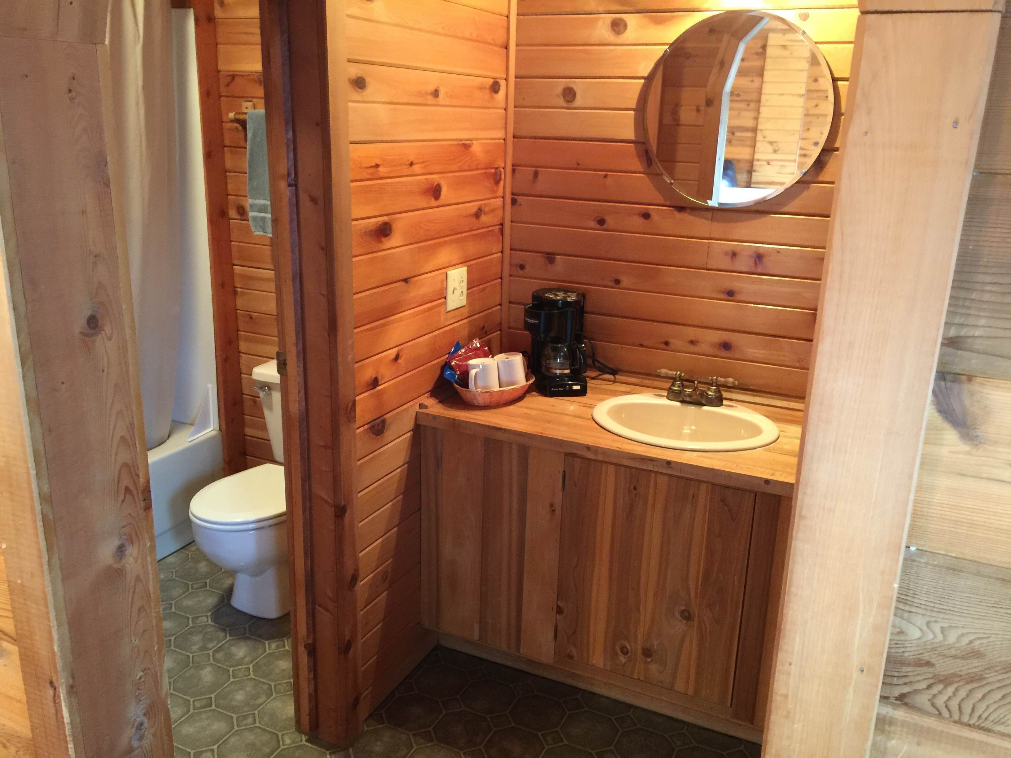 A wooden bathroom with a white sink, a round mirror, and a coffee maker.