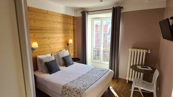 A bedroom with a wooden headboard, a bed with a patterned bedspread, a bedside table with a lamp, a chair, and a view of buildings through a window.