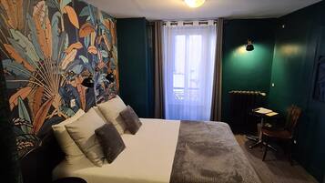 A hotel room with a bed, a small table, a chair, a wall with a tropical leaf pattern, and a window with curtains.