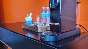 A coffee machine with a tray of cups, utensils, and water bottles on a black table against an orange wall.