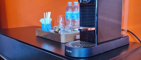 A coffee machine with a tray of cups, utensils, and water bottles on a black table against an orange wall.