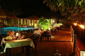 An outdoor dining area with tables set for a meal, a pool, and a tropical plant arrangement.