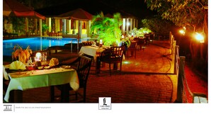 An outdoor dining area with tables set for a meal, a pool, and a building in the background.