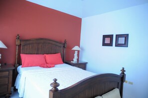A neatly made bed with a wooden headboard, two bedside tables with lamps, and two framed pictures on the wall. - Country Club Mangowalk Townhouse (Montego Bay)