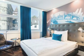 A hotel room with a large bed, a chair, a small table, and a view of a cityscape through the window.