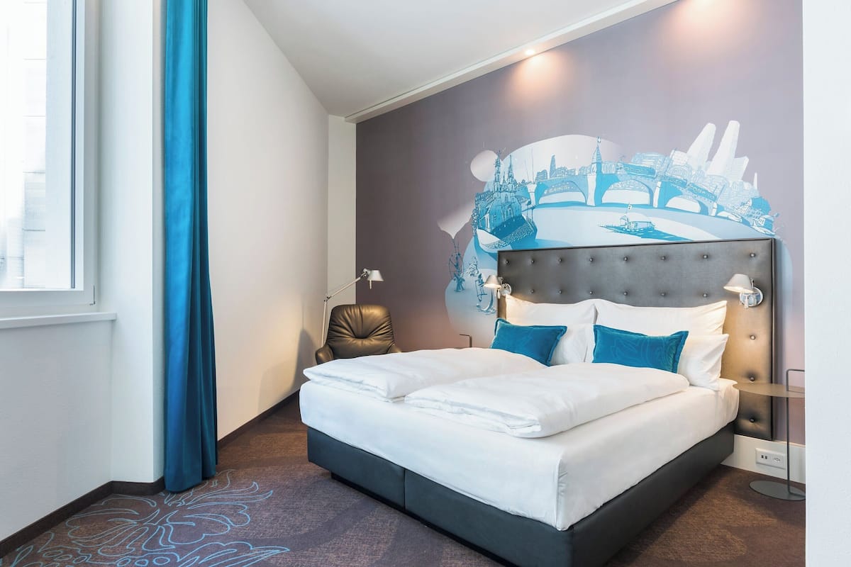 A modern hotel room with a large bed, a cityscape mural on the wall, a window with blue curtains, and a leather chair.