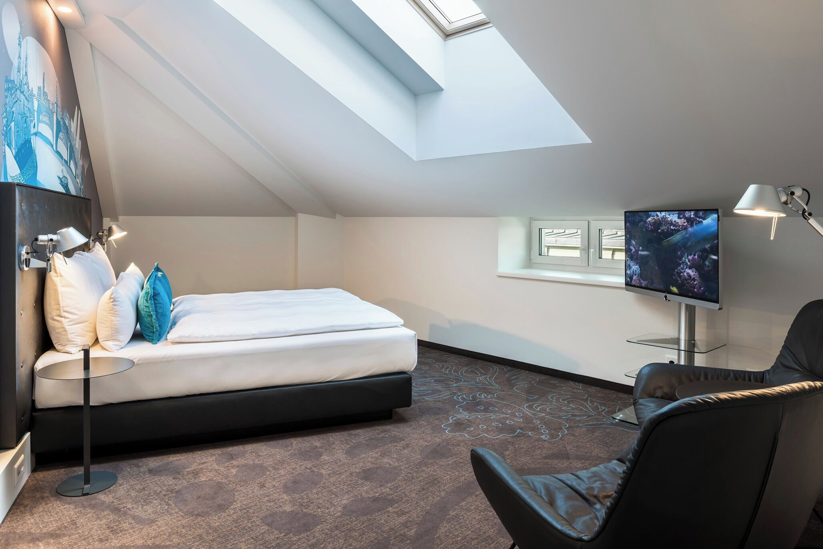 A modern hotel room with a skylight, a bed with white linens, a flat-screen TV, a chair, and a desk lamp.