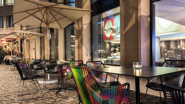 An outdoor dining area with tables and chairs, large umbrellas, and a view of the interior of a building. - Motel One Basel (Basel)