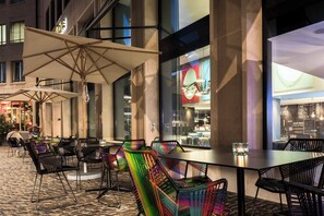 Un espace extérieur aménagé pour les repas, avec des tables et des chaises, de grands parasols et une vue sur l’intérieur d’un bâtiment.
