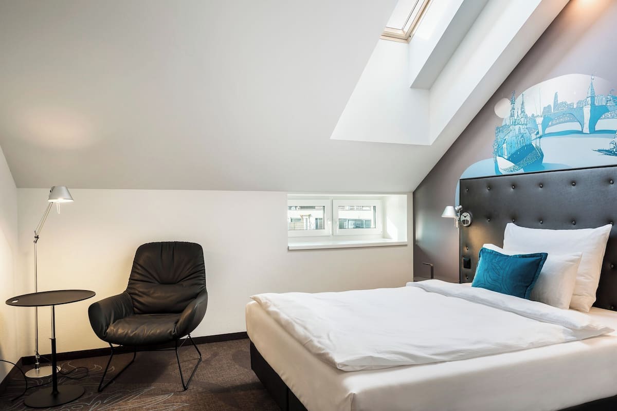 A hotel room with a bed, a chair, a lamp, and a window.