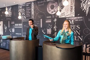 Two receptionists at a counter with a black wall featuring white text and graphics.