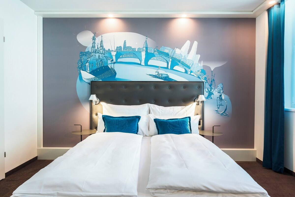 A hotel room with a bed, two bedside tables, a wall mural, and a window with curtains.