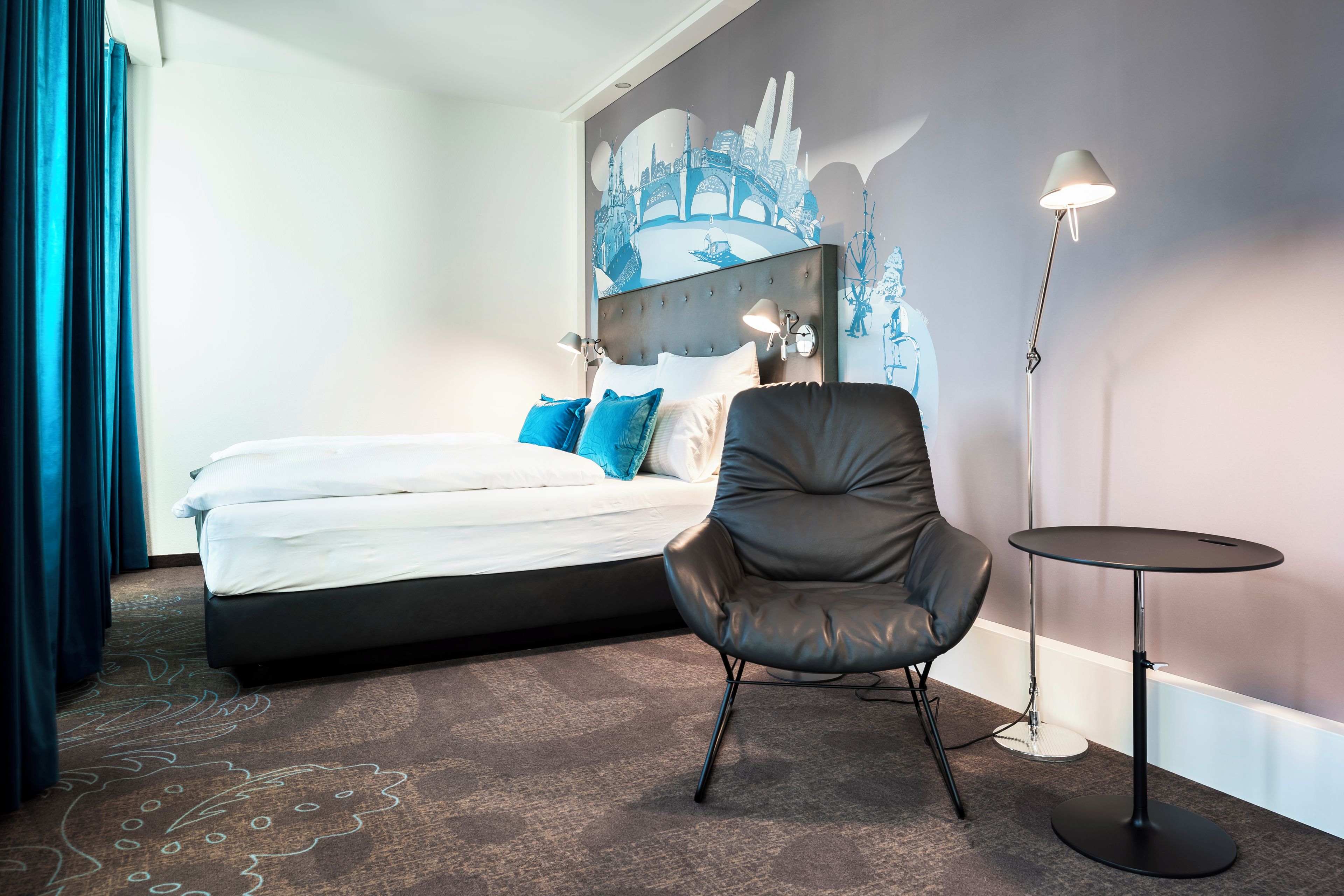 Habitación de hotel con cama, una silla, una mesita y un mural de un paisaje urbano en la pared.