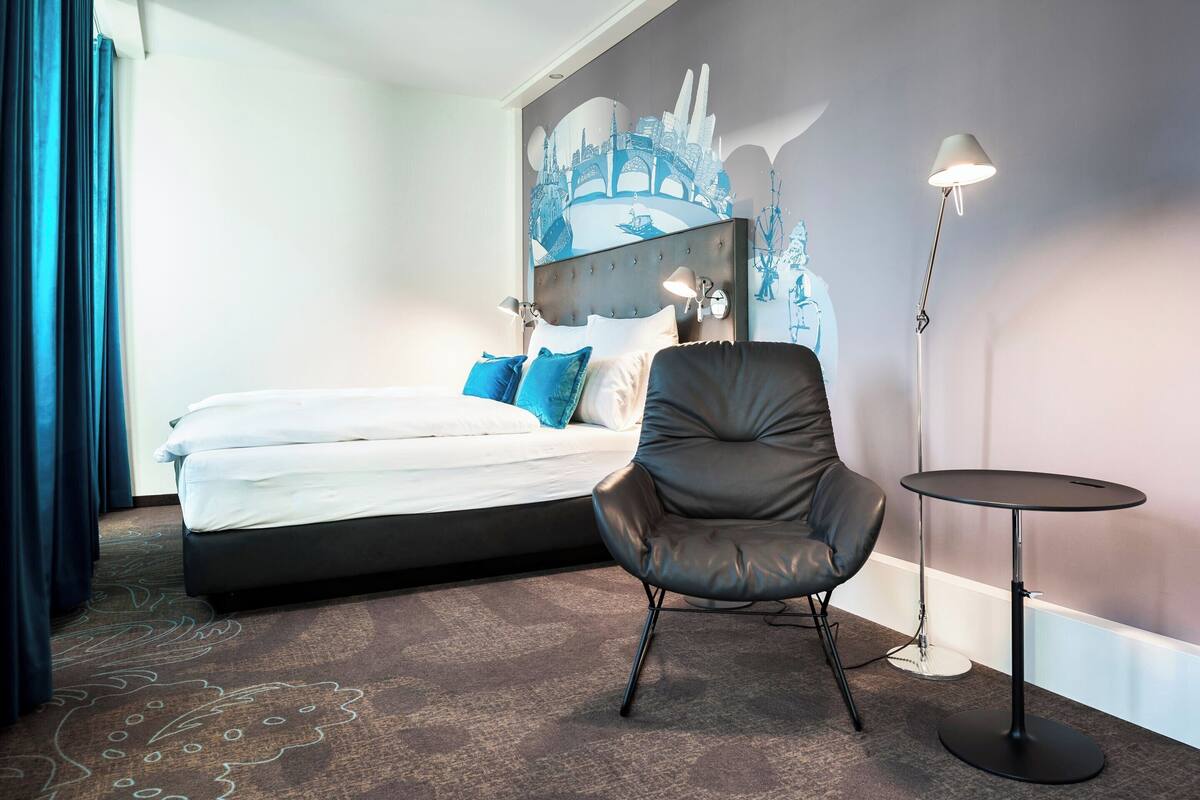 Une chambre d’hôtel moderne avec un grand lit, un fauteuil en cuir, une petite table ronde et une fresque représentant un paysage urbain sur le mur.