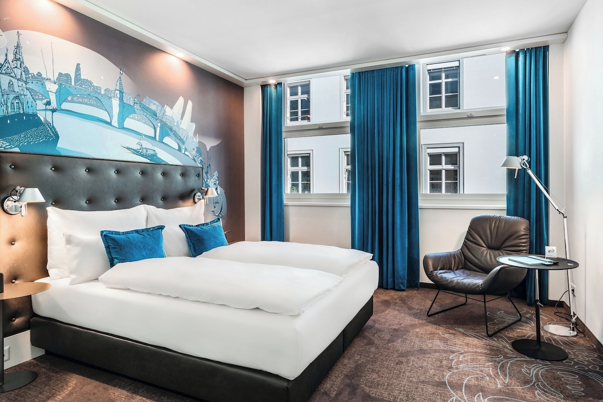 Habitació d'hotel amb un llit gran, una cadira, escriptori, làmpada i finestra amb cortines.
