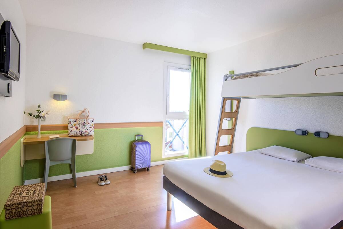 Una habitación de hotel con una cama individual, un escritorio con silla, una maleta, un sombrero y un taburete pequeño.