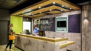 A reception desk with a wooden counter, a large window, and a wall with abstract art. - ibis budget Cannes Mougins (Mougins)