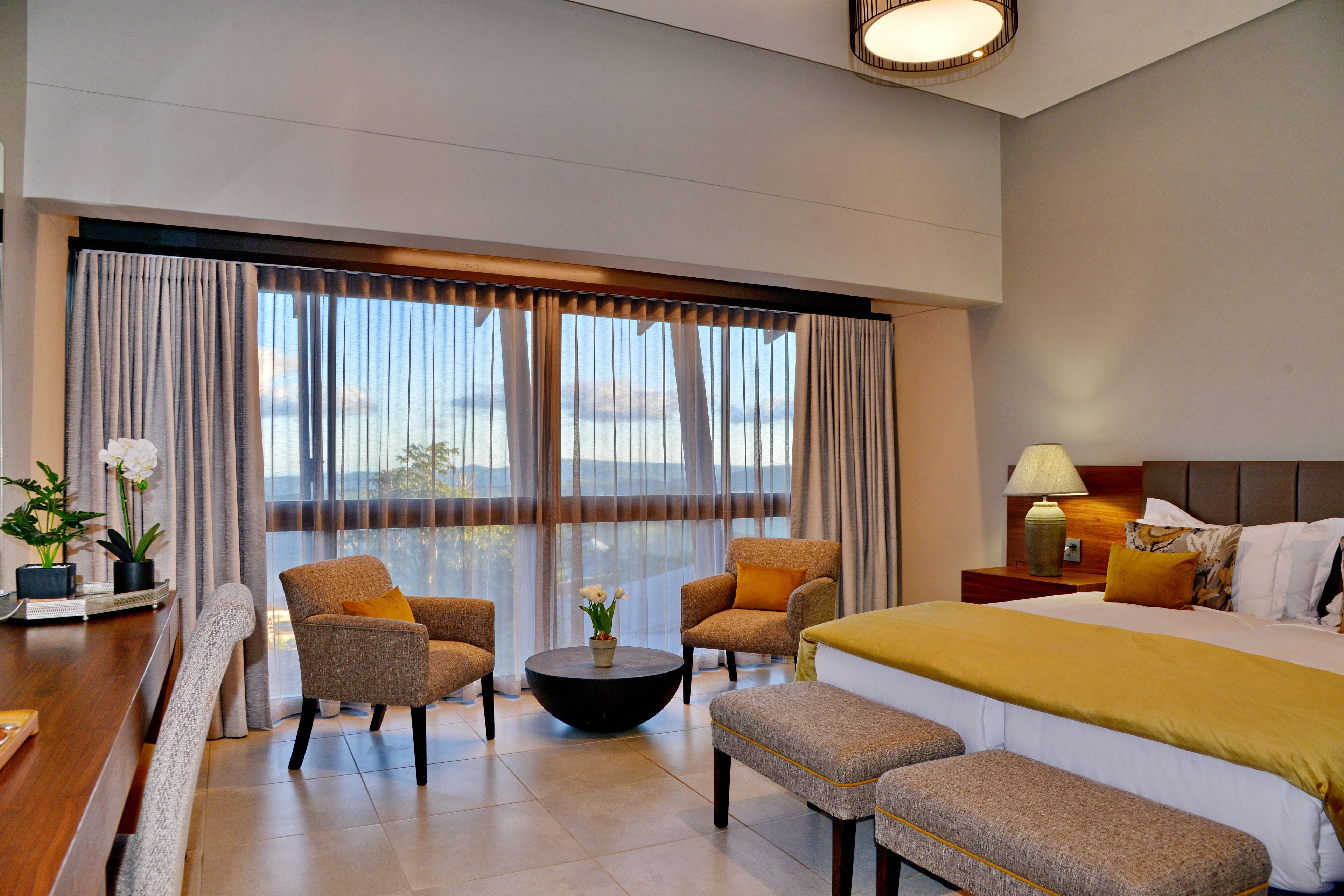 A hotel room with a large bed, two armchairs, a round coffee table, and a view of the outdoors through large windows.