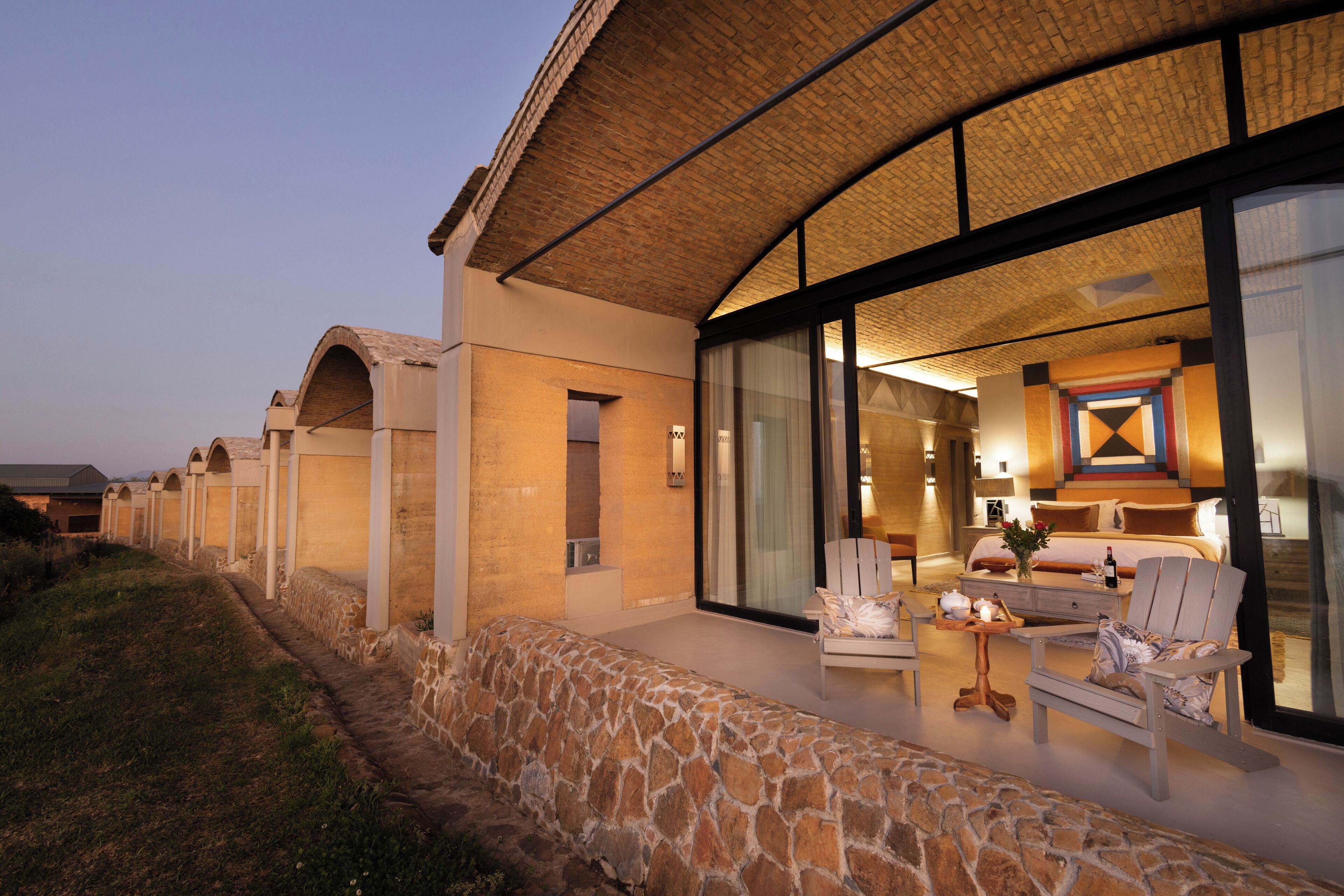 A modern hotel with a stone exterior, large windows, and a well-lit interior with a bed, chairs, and a dining area.
