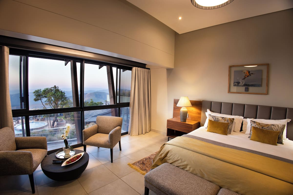 Una habitación de hotel moderna con un ventanal grande, una cama con ropa de cama dorada, una mesita de noche con lámpara y una silla junto a la ventana.
