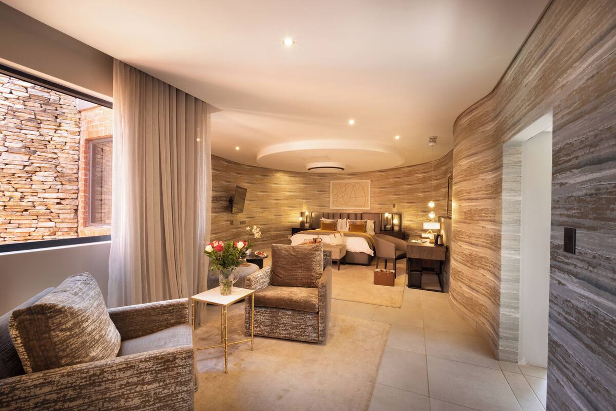 Una habitación de hotel moderna con pared de piedra, una cama grande, una zona de estar con una silla y una mesa, y vistas a través de una ventana.