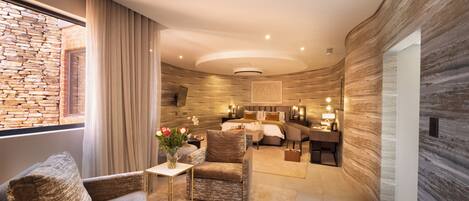 A modern hotel room with a stone wall, a large bed, a sitting area with a chair and table, and a view through a window.