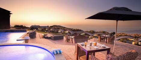 Zona junto a la piscina con sillones de descanso, una mesa y una sombrilla, con vistas a un paisaje pintoresco al atardecer.