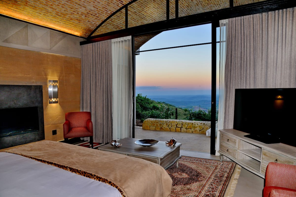 Una habitación de hotel moderna con una cama grande, un televisor, un sillón rojo, una mesa de comedor y una vista panorámica a través del ventanal.