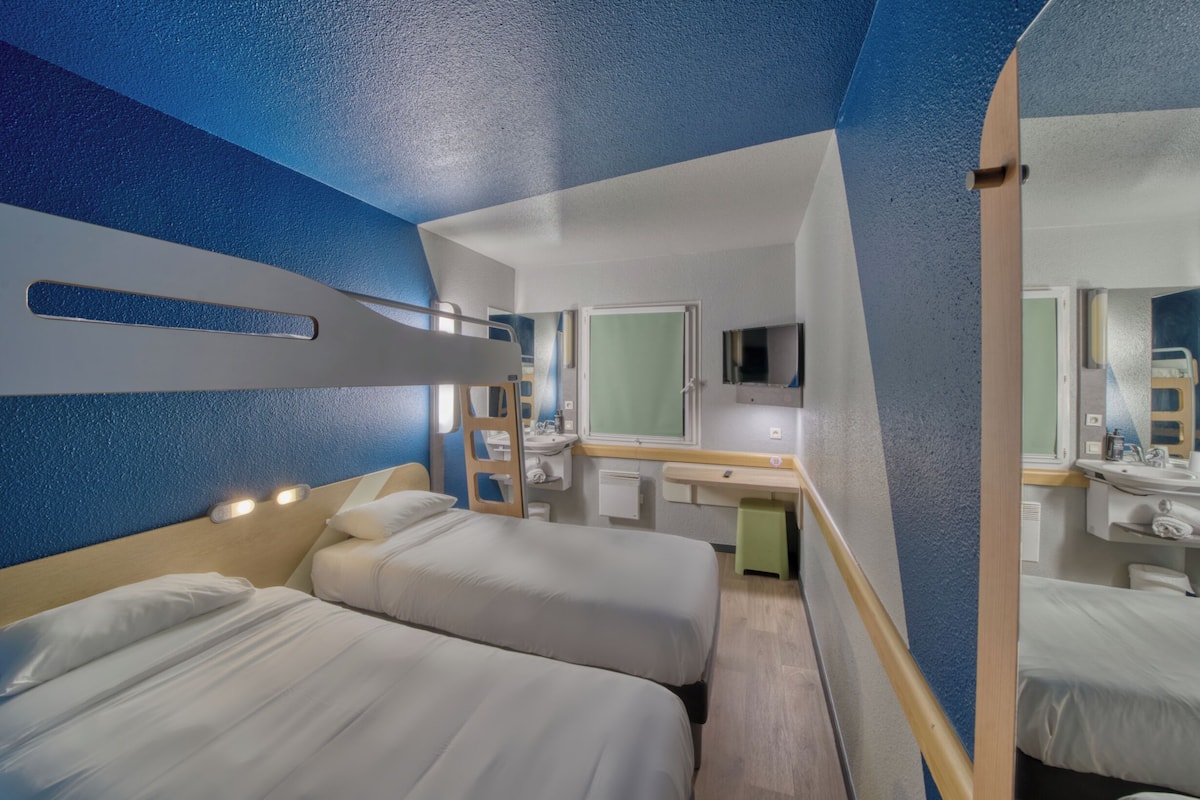 Quarto de hotel moderno com duas camas, um banheiro pequeno e uma área de trabalho.