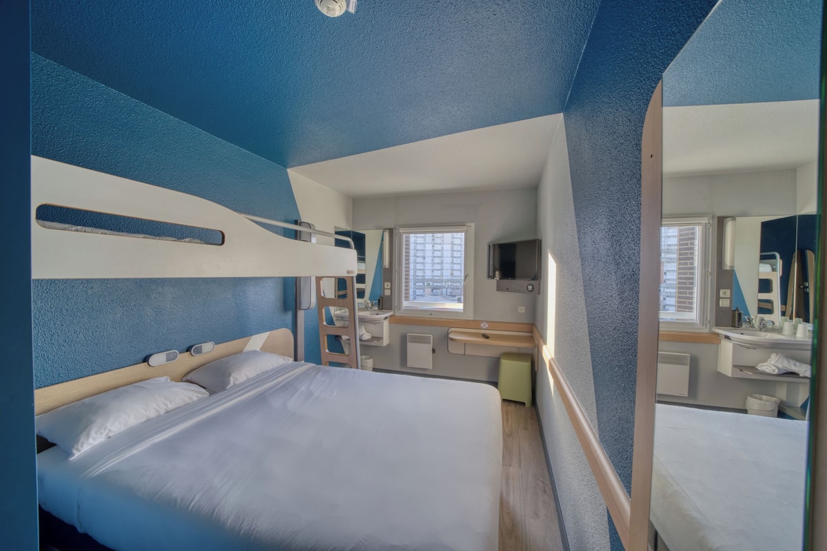 Quarto de hotel compacto com beliche, banheiro pequeno e janela que permite entrada de luz natural.