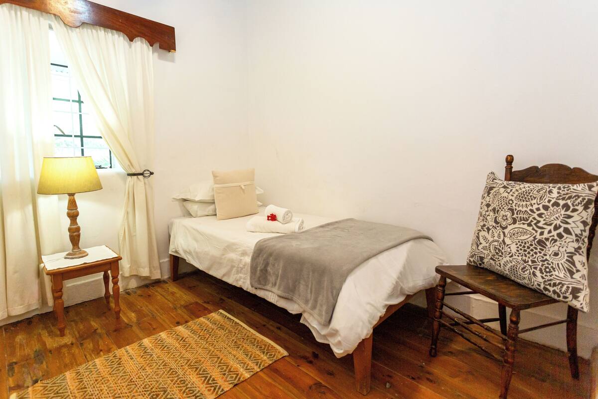 Una cama individual con edredón blanco, una mesita de noche con lámpara, una silla estampada y una alfombra con diseño.