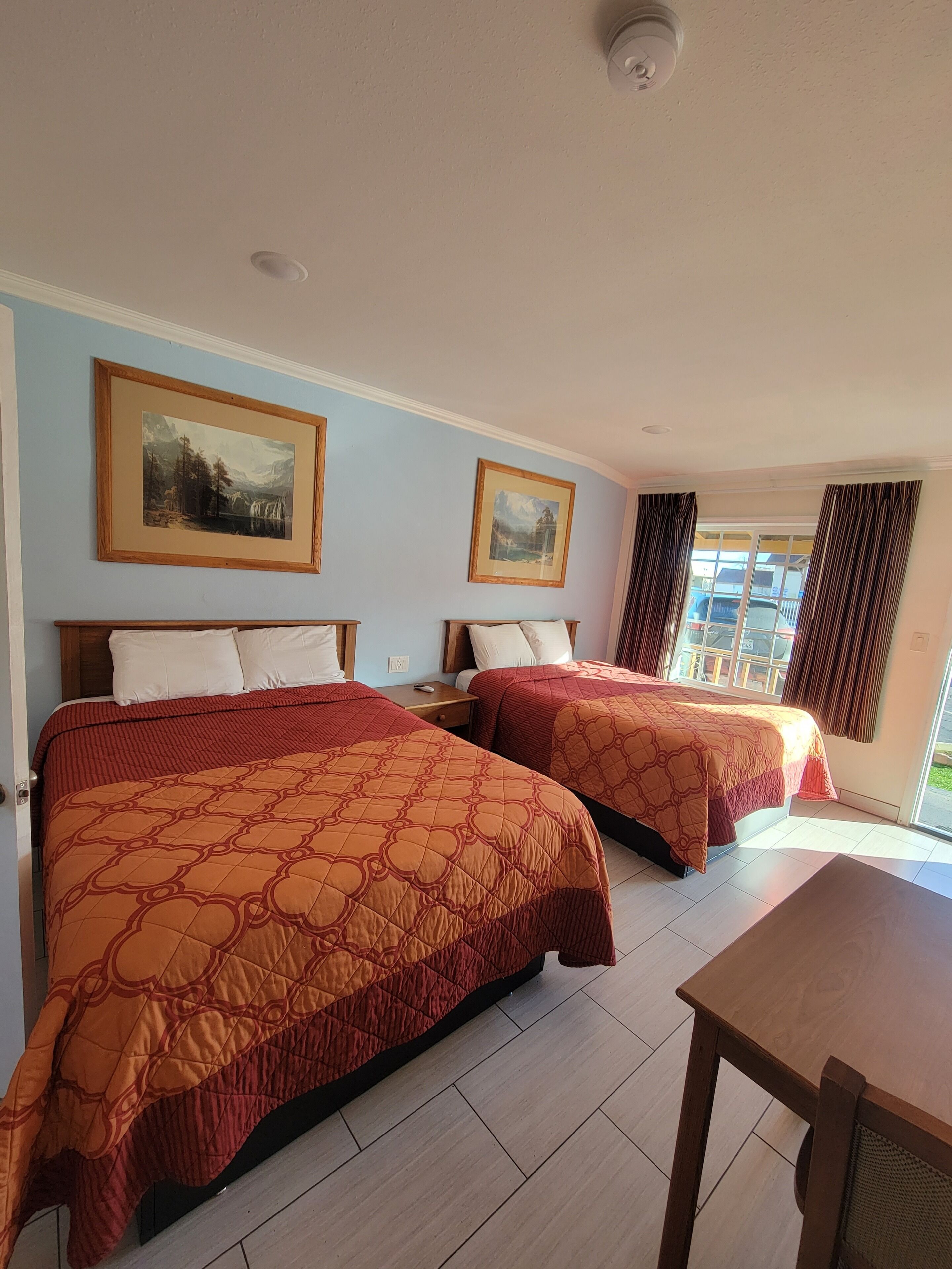 A hotel room with two beds, a wooden nightstand, a table, and two framed pictures on the wall.
