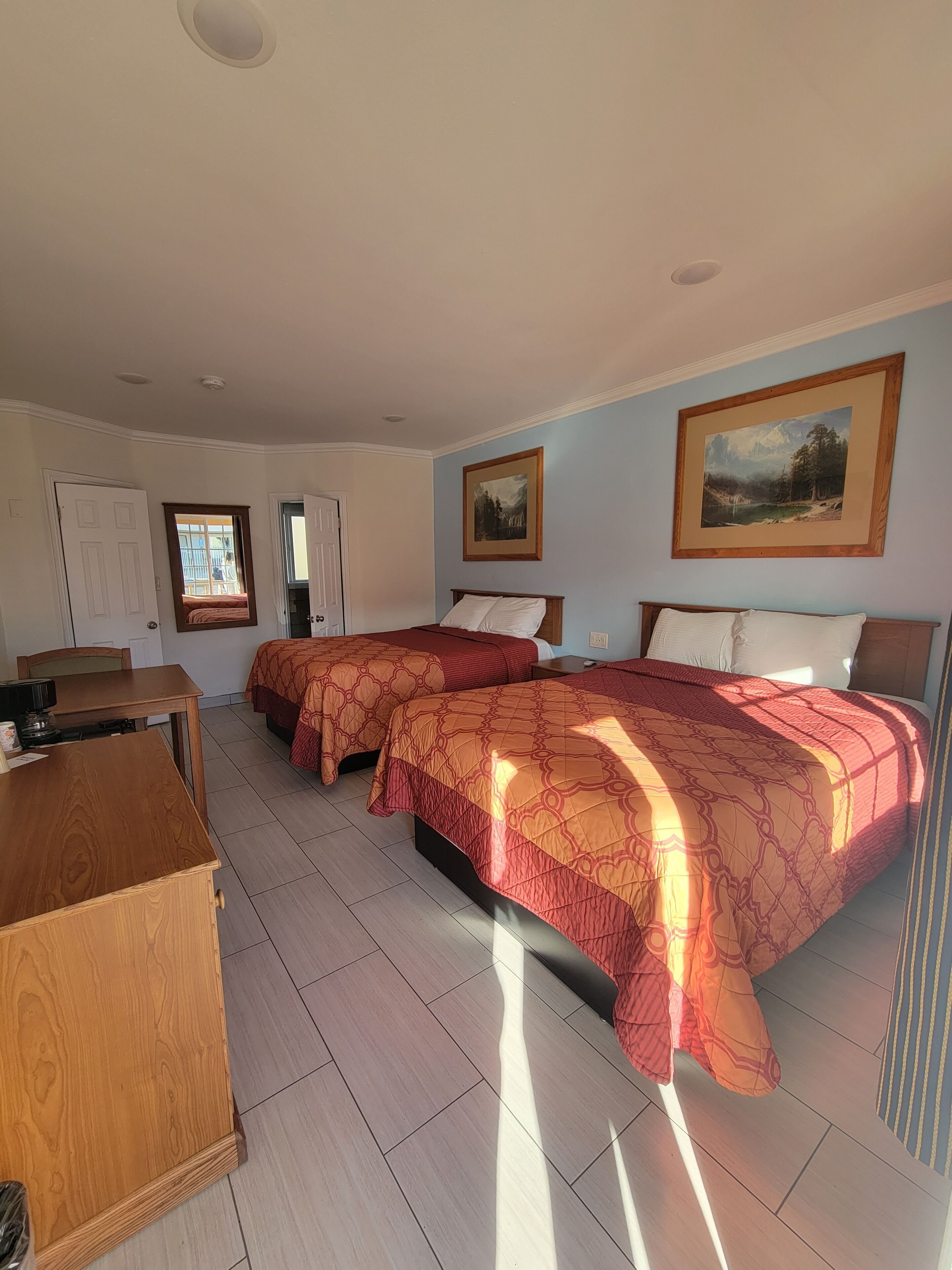 A hotel room with two beds, a desk, and a chair. There are two paintings on the walls and a window with a view of the outdoors.