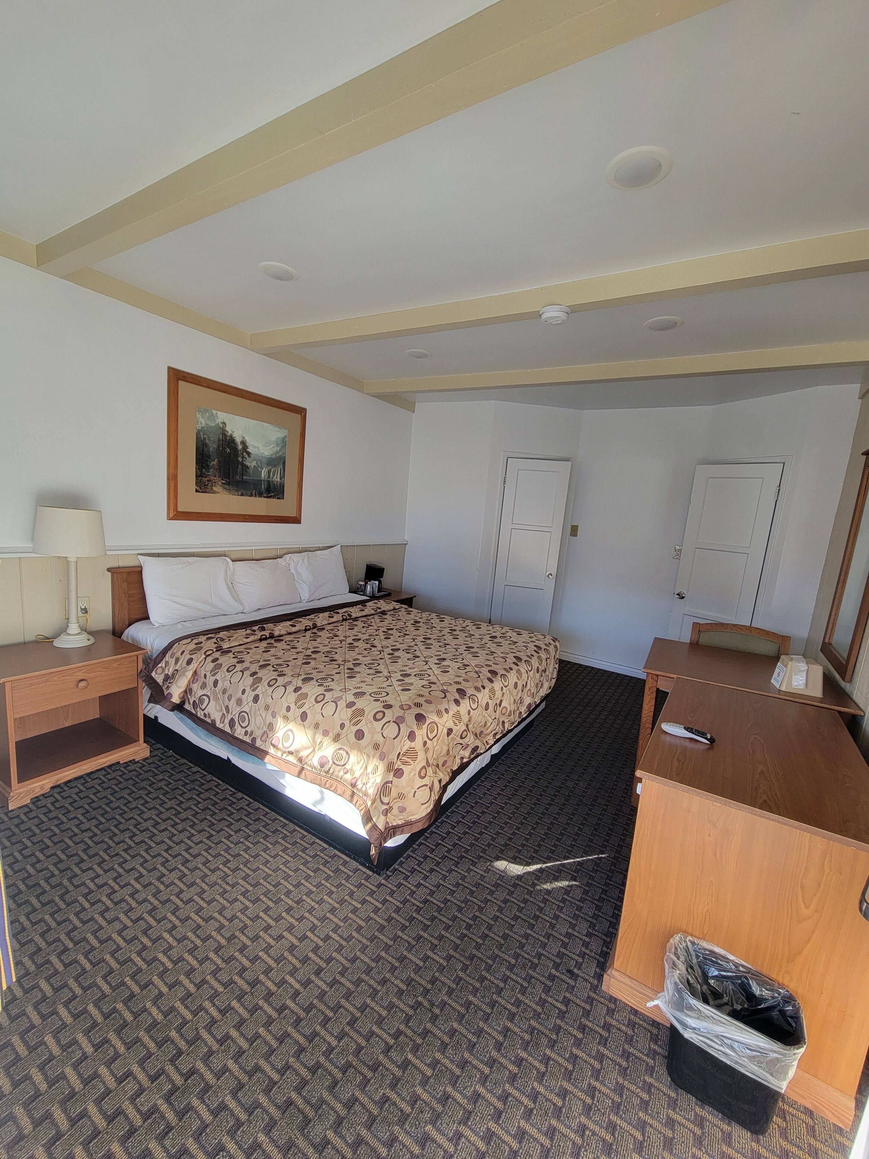 A hotel room with a large bed, a desk, a chair, a lamp, a painting on the wall, and a trash bin.