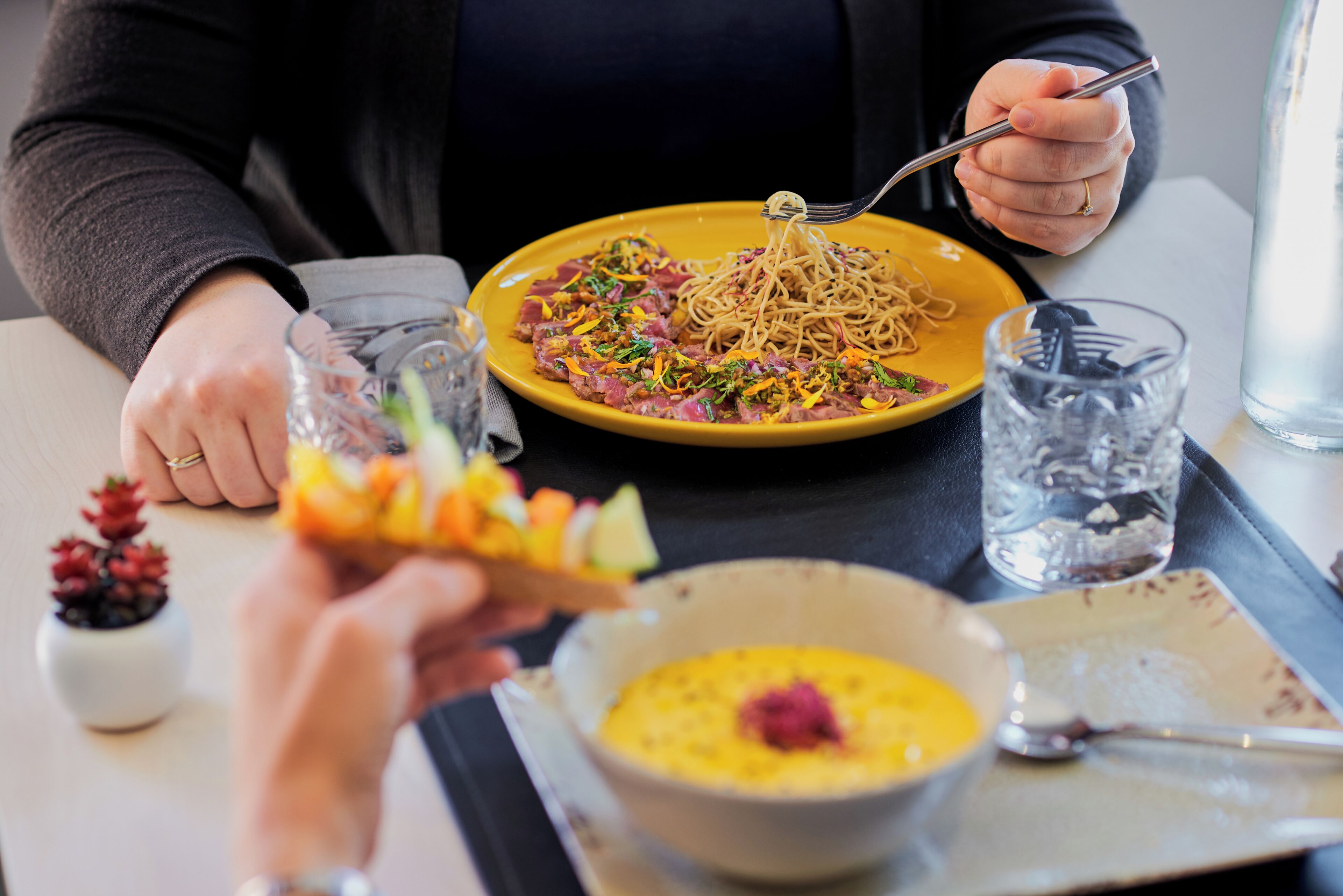 Une table à manger avec une assiette de spaghettis, un bol de soupe et un verre d’eau.