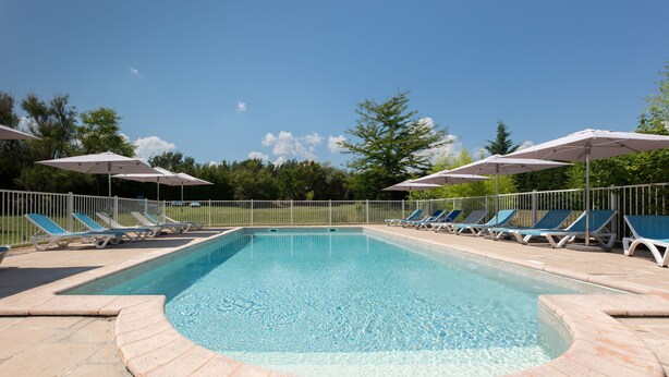 A rectangular swimming pool with a tiled edge, surrounded by a white fence. There are multiple lounge chairs and umbrellas along the poolside.