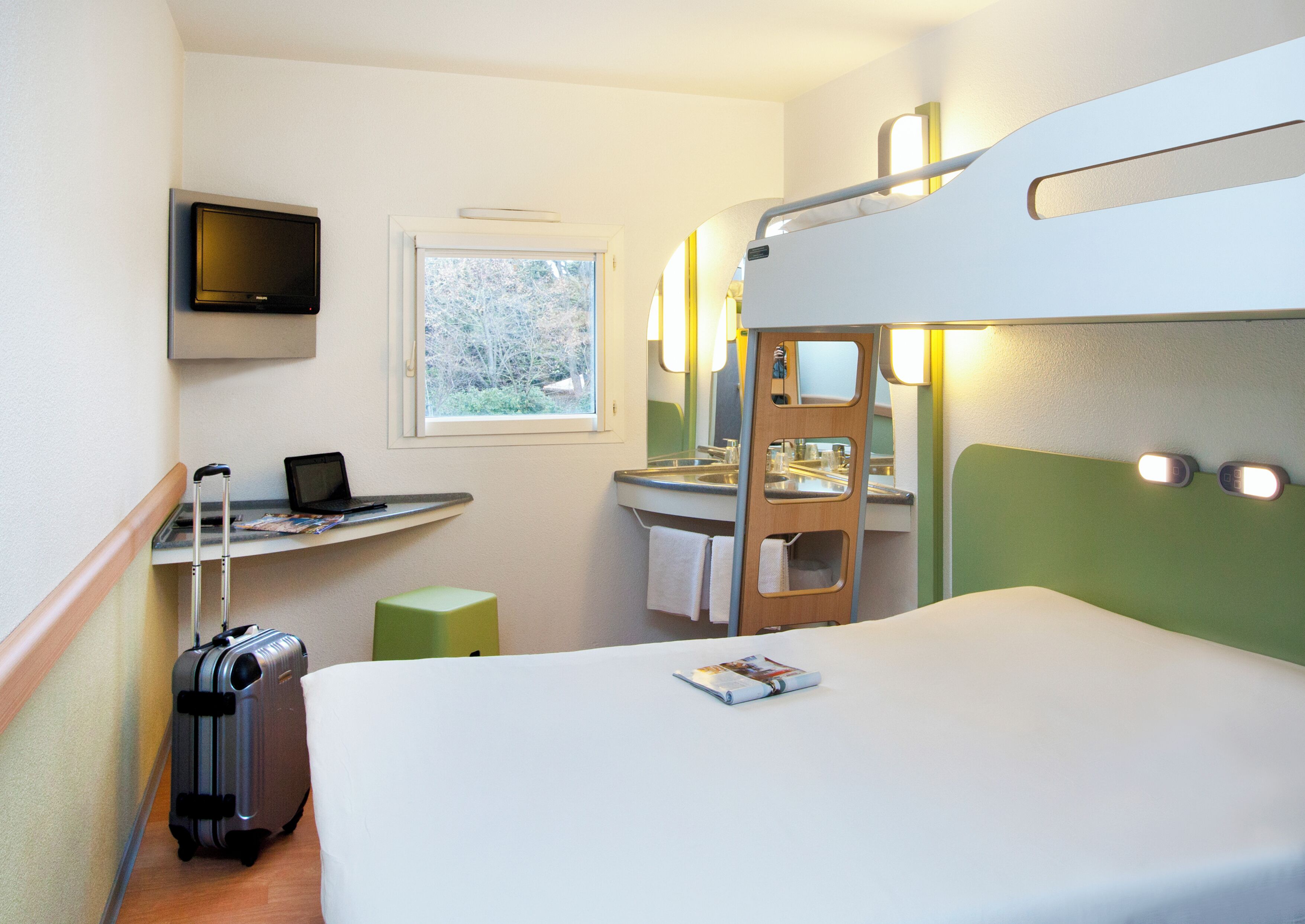 Et kompakt hotelværelse med køjeseng, et skrivebord med en bærbar computer, en kuffert og et vindue med udsigt til træer.