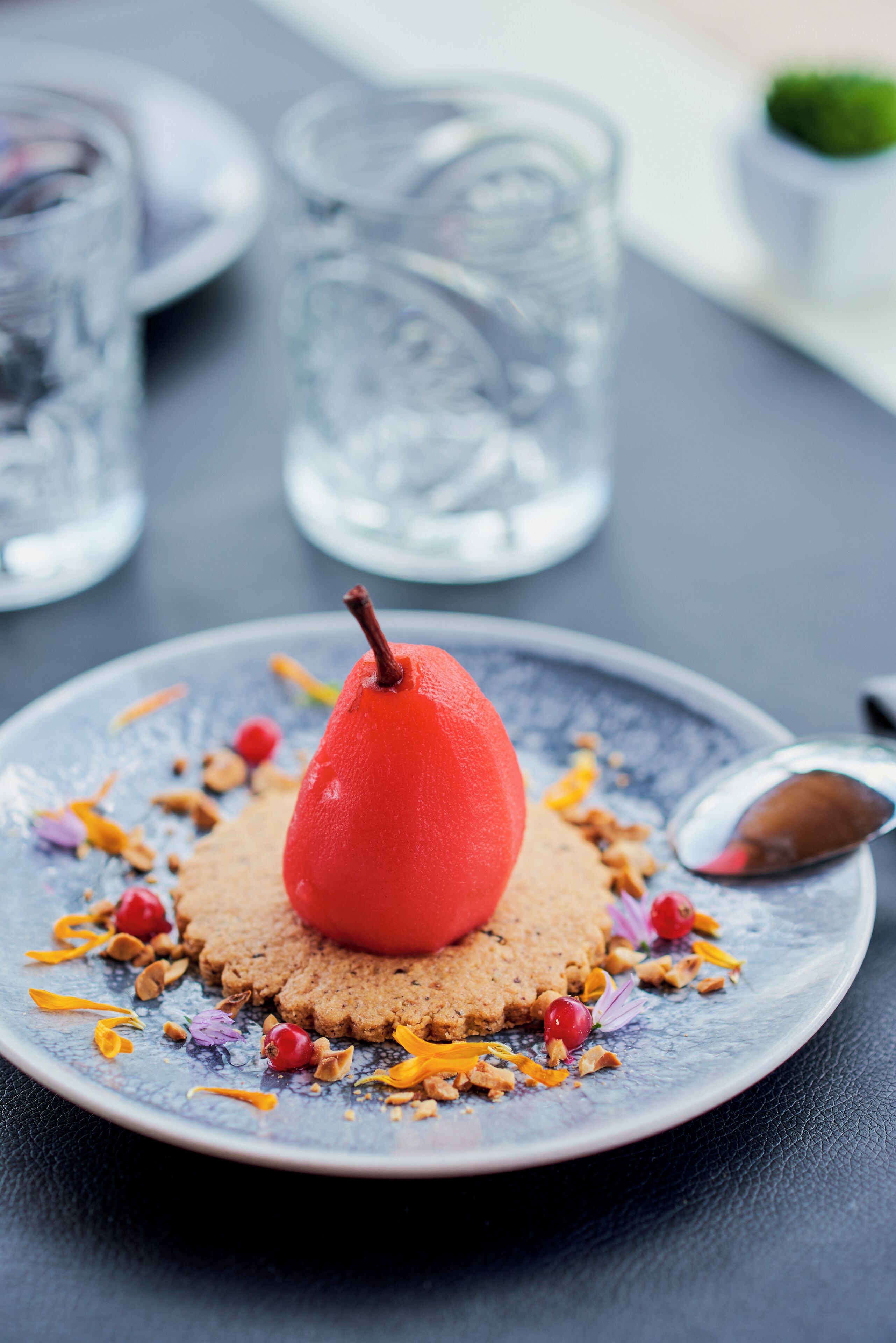 Un plato de postre con una pera roja encima, rodeado de nueces esparcidas y flores secas.