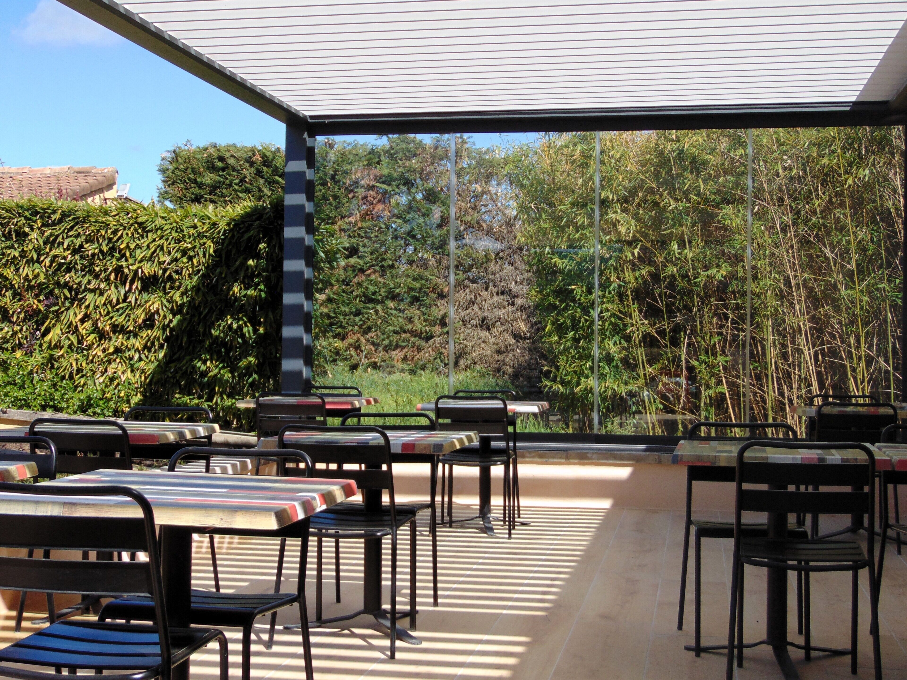 An outdoor patio with tables and chairs, surrounded by dense foliage and a clear sky.